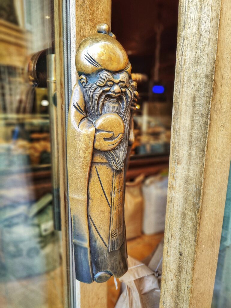 A bronze sculpture of a smiling monk holding a loaf of bread serves as a door handle at Farinoman Fou in Aix-en-Provence, France.